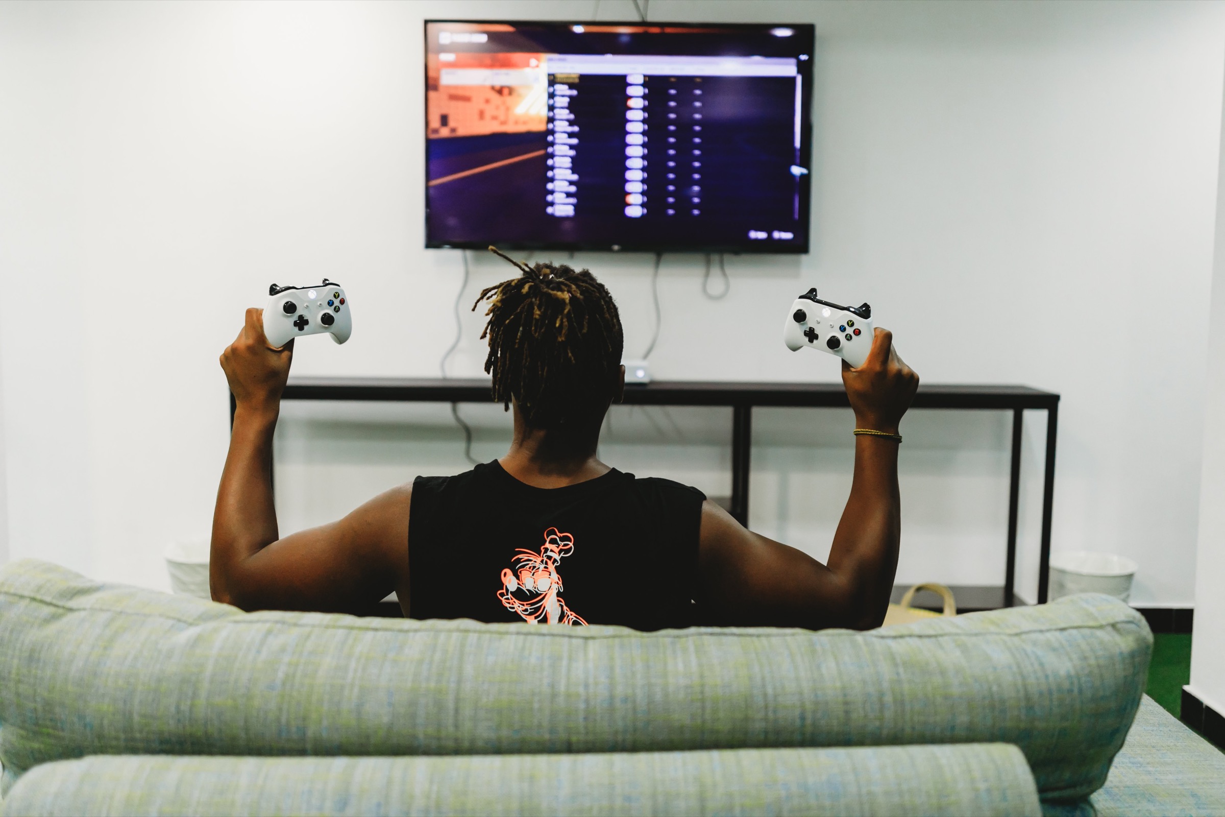 Person on a couch holding two game controllers in front of a wall-mounted TV